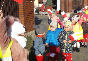 Grupa maluszków z flagami Polski spaceruje w pobliżu przedszkola