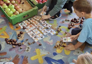 Dzieci rozwiązują na dywanie zagadki związane z jabłkami.