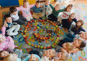 Dzieci siedzą na dywanie z ułożoną z jabłek uśmiechniętą buźką.
